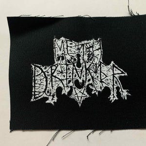 Może przedstawiać: Czarna prostokątna naszywka z białym tekstem i grafiką przypominającą nietoperza. Tekst brzmi "METAL DRINKER" w stylizowanej czcionce, z pentagramem na dole. Naszywka ma postrzępione krawędzie.