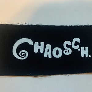 Puede incluir: Parche blanco bordado sobre tela negra con el texto "CHAOS C.H."