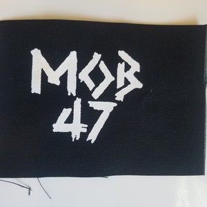 以下が含まれることがあります： 黒い長方形のパッチに、白い文字で「MOB 47」とプリントされています。「47」の数字は文字の下にあります。パッチは粗く、未完成の縁があります。