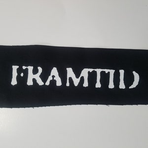 Peut inclure: Un patch rectangulaire noir avec le mot "FRAMTID" en blanc. Les lettres majuscules sont stylisées. Le patch a des bords bruts, indiquant qu'il est probablement en tissu. Le texte est l'élément principal.