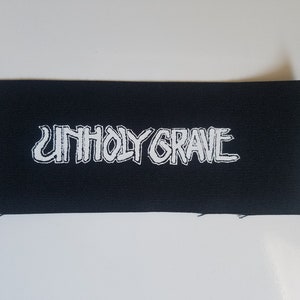May include: Black rectangular patch with the white text "UNHOLY GRAVE". The text has a distressed, gothic font style. The patch is made of a woven fabric with a raw edge.