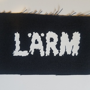 May include: Black rectangular patch with the word "L'ARM" in white, stylized, block letters. The edges of the patch are frayed, with loose threads visible. The patch is likely made of fabric.