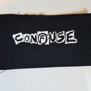 May include: White "CONFUSE" text on a black fabric patch. The letters are in a distressed, cut-out style.