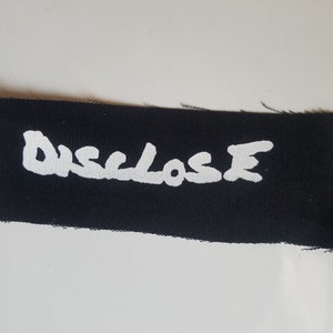 May include: Black fabric patch with the word "DISCLOSE" written in white paint.