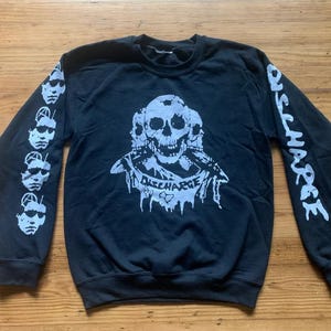 Könnte beinhalten: Schwarzer Langarm-Pullover mit einem weißen Aufdruck von drei Totenköpfen und dem Wort "DISCHARGE" auf der Vorderseite. Die Ärmel haben einen weißen Aufdruck des Wortes "DISCHARGE", der sich über die gesamte Länge des Ärmels wiederholt.