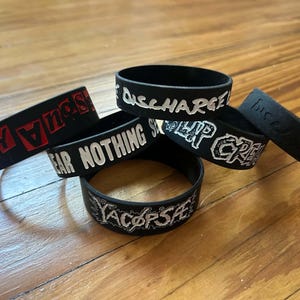 May include: A collection of five black silicone wristbands featuring various white and red printed text designs. The bands display words including DISCHARGE, FEAR NOTHING, LIP CREAM, and YACOPSAE in distinct fonts, arranged in a small cluster.