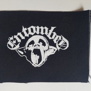 Könnte beinhalten: Weiß gestickter Patch auf schwarzem Stoff mit dem Text "Entombed" und einer Grafik eines schreienden Schädels.