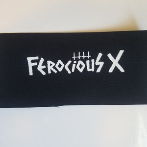 Pode incluir: Remendo de tecido preto com texto bordado branco que diz "FEROCIOUS X" com quatro sinais de mais acima da palavra "FEROCIOUS".