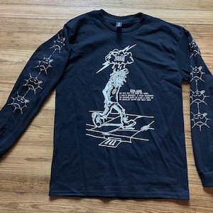 May include: Black long-sleeved shirt featuring a white graphic of a person with a cloud and lightning bolt above their head. The shirt has the text "LIFE LOCK". The sleeves have a series of white sun-like designs.