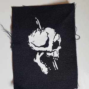 Könnte beinhalten: Ein schwarzer Stoff-Patch mit einem weißen Totenkopf- und Knochen-Design. Der Schädel ist nach rechts gerichtet und hat eine große, hervorstehende Augenhöhle.