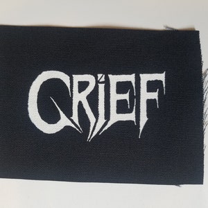 Pode incluir: Patch de tecido preto com a palavra "GRIEF" em branco, em letras estilizadas.