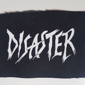 以下が含まれることがあります： 黒い布のパッチに、スタイリッシュなフォントで「DISASTER」という文字が白で印刷されています