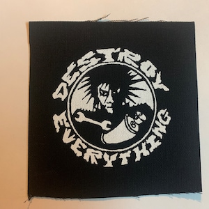 Op de afbeelding: Zwart-witte geborduurde patch met een cirkelvormig ontwerp. De cirkel toont een figuur met stekelig haar die een moersleutel en een blikje vasthoudt. De tekst "DESTROY EVERYTHING" staat rond de cirkel.