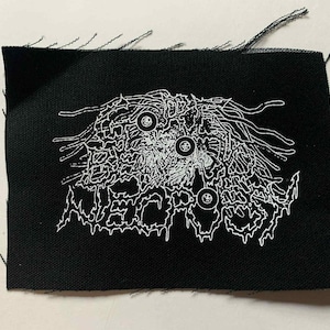 May include: Black fabric patch with a white graphic design. The design features a detailed, abstract illustration with the word "NECROPSY" in a stylized font. The patch has frayed edges.