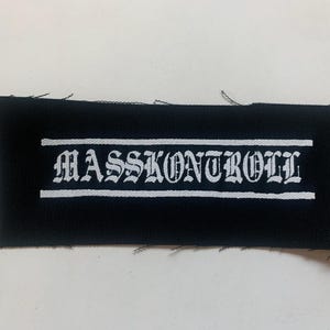 以下が含まれることがあります： ゴシック体で「MASSKONTROLL」と書かれた白いテキストが施された黒い布のパッチ