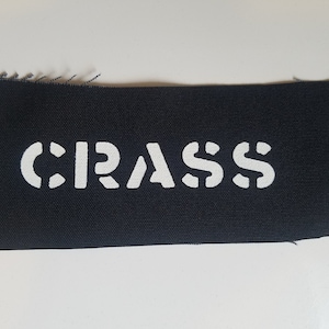 Peut inclure: Un patch en tissu noir avec le mot "CRASS" imprimé en blanc.