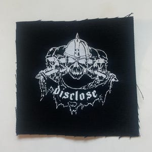 May include: Black square patch with a white graphic of three skulls wearing helmets, with the word "disclose" in a gothic font. The patch is made of fabric and has a raw edge.