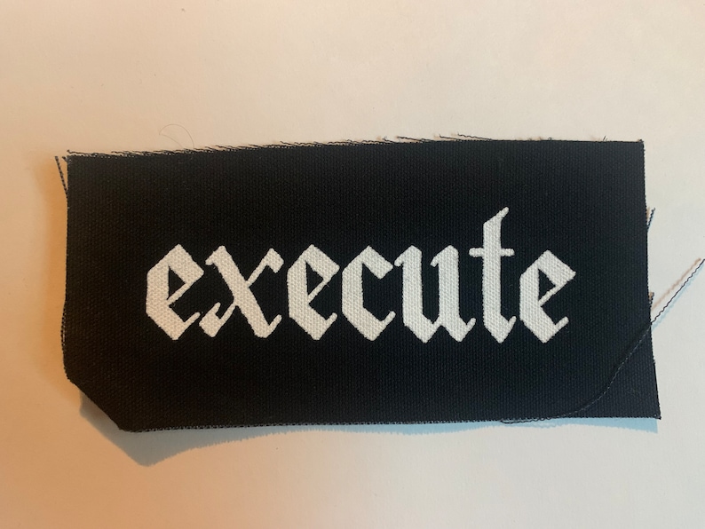 K&ouml;nnte beinhalten: Schwarzer Stoff-Patch mit dem Wort "execute" in wei&szlig;er gotischer Schrift.
