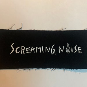 May include: Black fabric patch with white embroidered text that reads "SCREAMING NOISE" and a simple white drawing of a face with a single strand of hair.