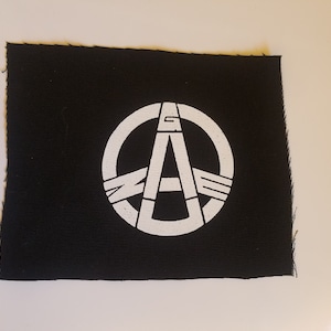 May include: Black square patch with a white graphic design. The design features a circle with the letters "G", "A", and "E" inside. The patch has a raw edge.