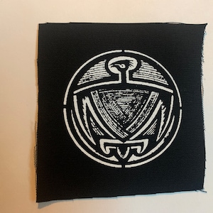 May include: White embroidered patch on black fabric featuring a stylized eagle design inside a circle.