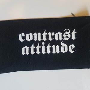 May include: A black rectangular patch featuring the words "contrast attitude" in white gothic font. The patch is made of fabric with frayed edges.