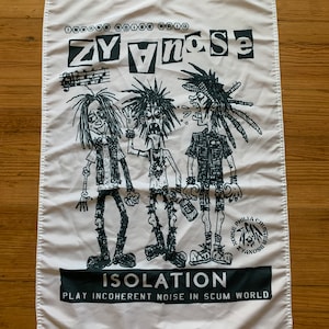 May include: A white flag with black text and illustrations. The text reads "ZY yno Se" and "ISOLATION PLAY INCOHERENT NOISE IN SCUM WORLD". The illustrations are of three cartoon figures with spiky hair and clothing. The flag has a black and white logo with the text "NOISE PHILLA CRUSTED ZYANOSE".