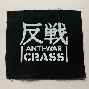 Może przedstawiać: Czarna kwadratowa naszywka z białym tekstem. Górna linia zawiera dwa japońskie znaki. Poniżej tekst brzmi „ANTI-WAR” i „CRASS” w czcionce szablonowej. Naszywka ma surową krawędź.