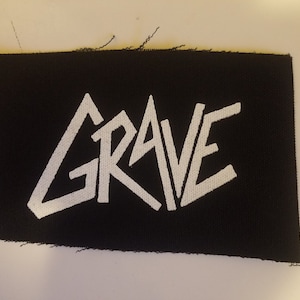 May include: A black fabric patch with the word "GRAVE" printed in white. The letters are stylized and angled.