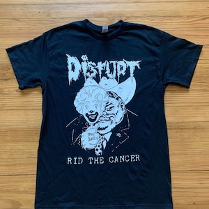 Puede incluir: Camiseta negra con un gráfico blanco de una persona con un sombrero de vaquero y una máscara de payaso. El texto "DISRUPT" y "RID THE CANCER" está impreso en la camiseta.
