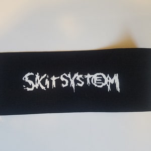 Op de afbeelding: Zwarte stoffen patch met witte geborduurde tekst die "SKITSYSYTEM" luidt.