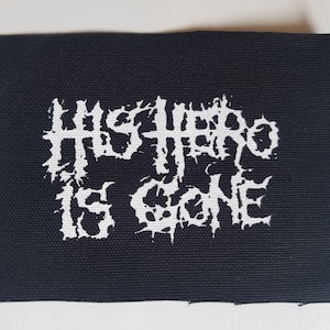 Op de afbeelding: Zwart-wit geborduurde patch met de tekst "The Hero Is Gone" in een gestileerd lettertype.