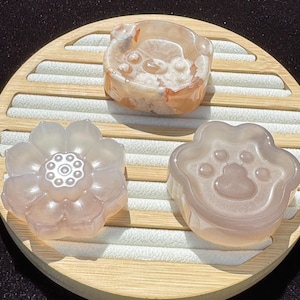 May include: Three carved stone objects on a wooden tray. The objects include a bear-shaped piece, a flower, and a paw print. The bear is orange and white, the flower and paw print are light pink. The tray has a light wood frame and white fabric.