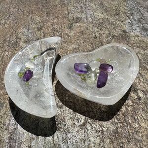 May include: Two clear quartz crystal dishes, one crescent-shaped and one heart-shaped, each filled with a collection of polished gemstones. The gemstones include amethyst, peridot, and clear quartz crystals. The dishes sit on a textured, gray surface.