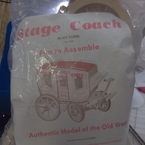 May include: A white plastic bag containing a wooden model kit of a stagecoach. The kit is labeled "Stage Coach" and "Authentic Model of the Old West". The kit is in the form of a model to be assembled. The bag is transparent and shows the model kit inside.