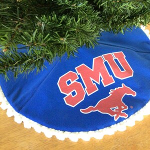 May include: A blue Christmas tree skirt with the letters "SMU" in red and a red mustang logo. The skirt has a white, scalloped edge and is partially covered by a green Christmas tree.