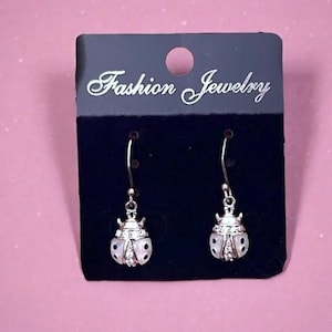 May include: A pair of silver-toned drop earrings shaped like ladybugs, featuring white enamel wings with black spots and small clear crystals along the center. The earrings are attached to a black display card with the text Fashion Jewelry.