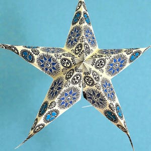 May include: A decorative paper star lantern with a blue and cream floral pattern. The star has five points and is suspended against a light blue background. The design features blue floral shapes and black outlines.