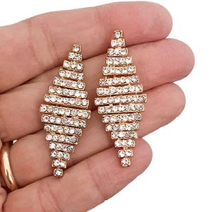 Puede incluir: Un par de pendientes geométricos dorados. Cada pendiente presenta un diseño en capas de cristales claros y brillantes engastados en un marco de metal dorado. Los pendientes tienen una forma larga y delgada.