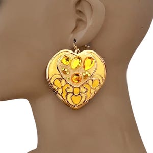 May include: Gold-toned heart-shaped earrings with yellow gemstones. The earrings feature a detailed design with a heart-within-a-heart pattern and are suspended from a simple hook. The gemstones are various shapes and sizes.