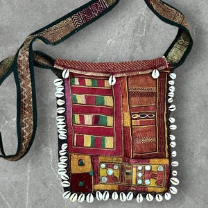 Könnte beinhalten: Eine farbenfrohe, quadratische Umhängetasche mit langem Riemen. Die Tasche hat ein Patchwork-Design mit roten, gelben und grünen geometrischen Mustern. Weiße Muscheln sind an den Rändern angenäht. Der Riemen ist dunkelgrün mit goldenen Akzenten.