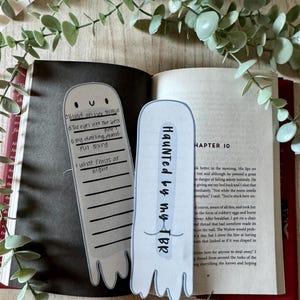 Puede incluir: Dos marcadores con forma de fantasma con texto, uno que dice "Haunted by my TBR" y el otro con notas manuscritas. Los marcadores están en un libro abierto con un lápiz cerca, con follaje verde en el fondo.