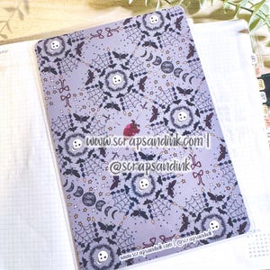 May include: A lavender planner cover with a repeating pattern of skulls, bats, spiderwebs, moons, and stars. The cover has the text "www.scrapsandink.com" and "@scrapsandink". The planner is open to a blank page.