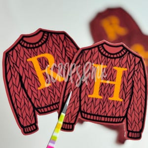 May include: Two red and black knit sweater stickers with yellow letters. The sweaters have the letters "R" and "H" on them.