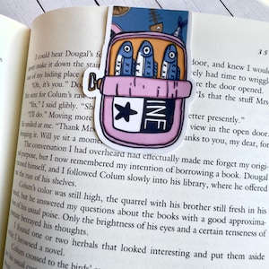 May include: A pink and blue bookmark with a cartoon illustration of a bucket with three fish inside. The bucket has a white star and the word "INE" written on it. The bookmark is placed on a book.