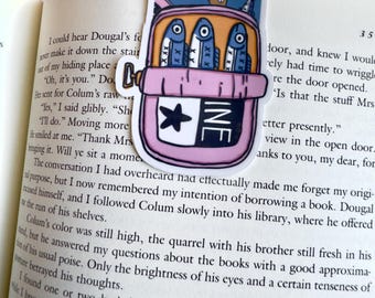 May include: A pink and blue bookmark with a cartoon illustration of a bucket with three fish inside. The bucket has a white star and the word "INE" written on it. The bookmark is placed on a book.