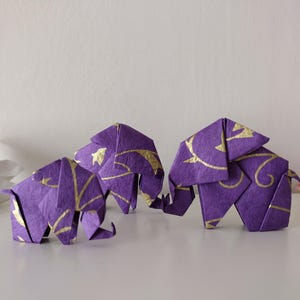 May include: Three origami elephants in shades of purple and pink, with gold decorative accents. The elephants are arranged on a white surface, with a white daisy in the background. The paper elephants are a unique decorative item.