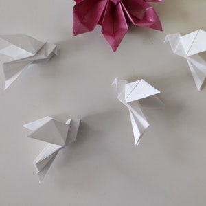 May include: Four white origami paper doves are arranged on a white surface. A single pink origami flower is in the centre of the image.