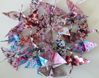 May include: A collection of origami paper cranes in various floral patterns, including pink, blue, and white. The cranes are folded from patterned paper and are arranged in a circular pattern.