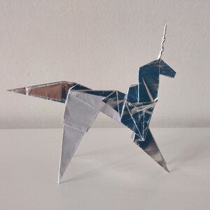 May include: A silver origami unicorn with a curled horn. The unicorn is standing on a white surface.